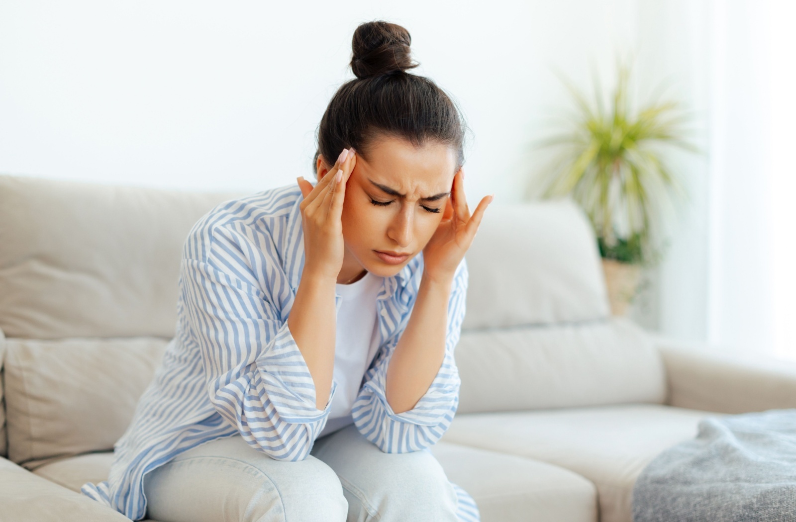 Woman sitting on a couch with her hands on her temples, eyes closed in discomfort.