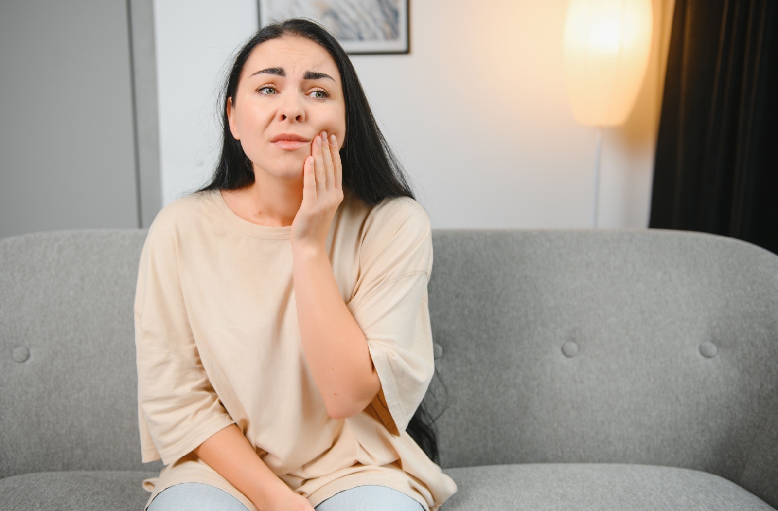 Woman sitting on a couch holding her jaw with a concerned expression.