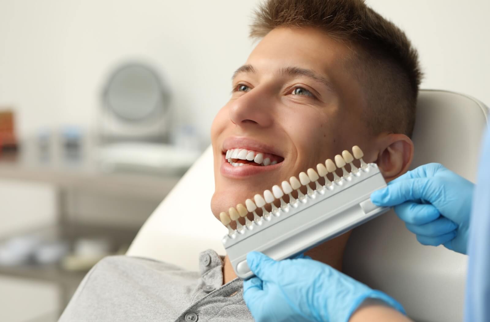 Young smiling man sits in the dental chair while off-camera dentist in blue gloves holds a set of veneers in varying colours to his teeth.