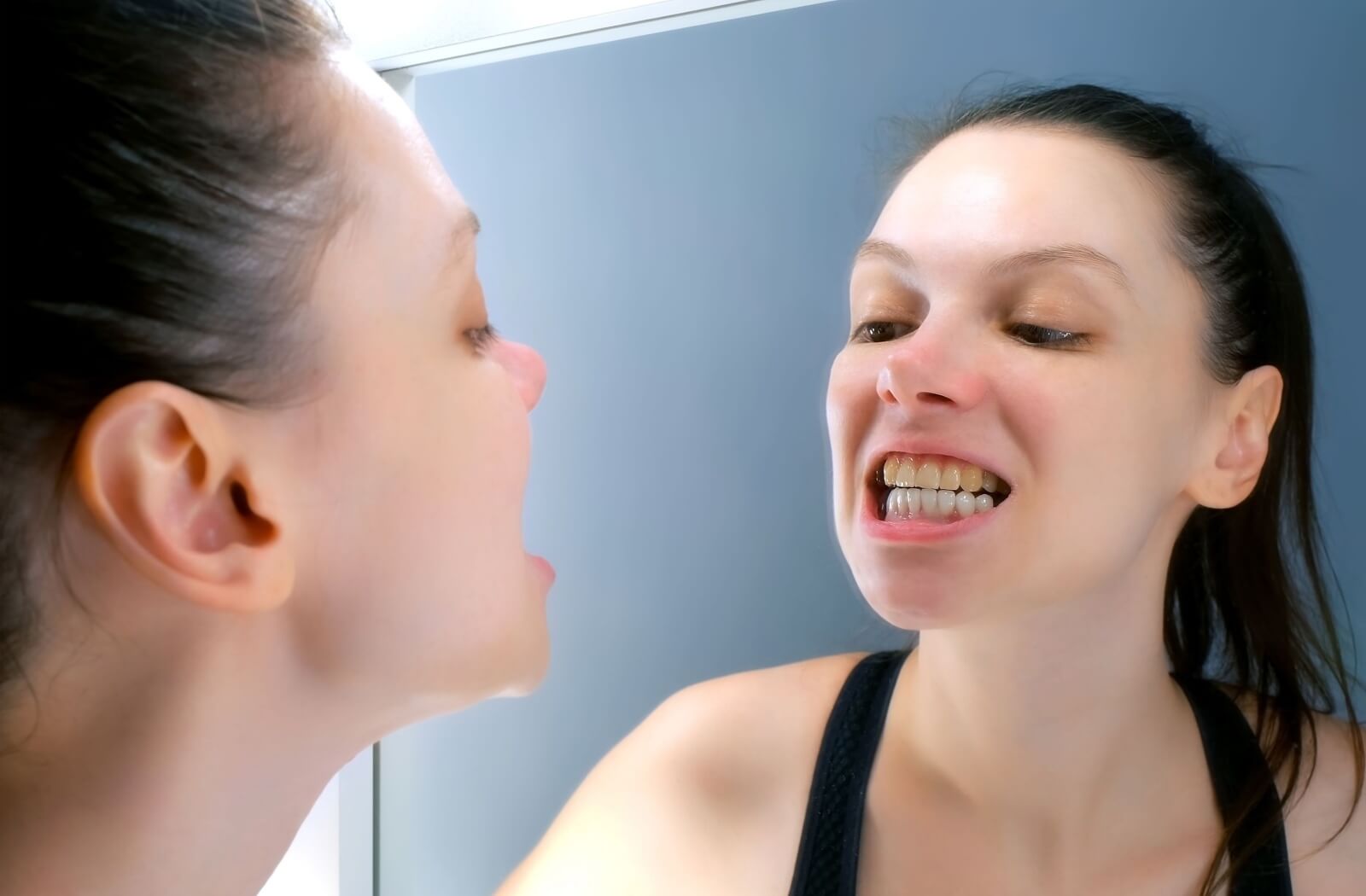 Young woman with dark ponytail looking at her teeth in the mirror. Top teeth have yellow staining.