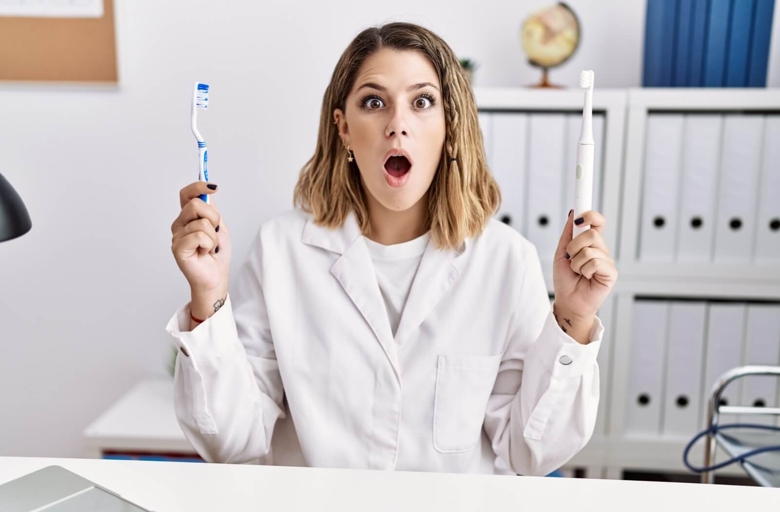 Female dentist wearing a white jacket in an examination office holding an electric toothbrush in one hand and a manual toothbrush in the other