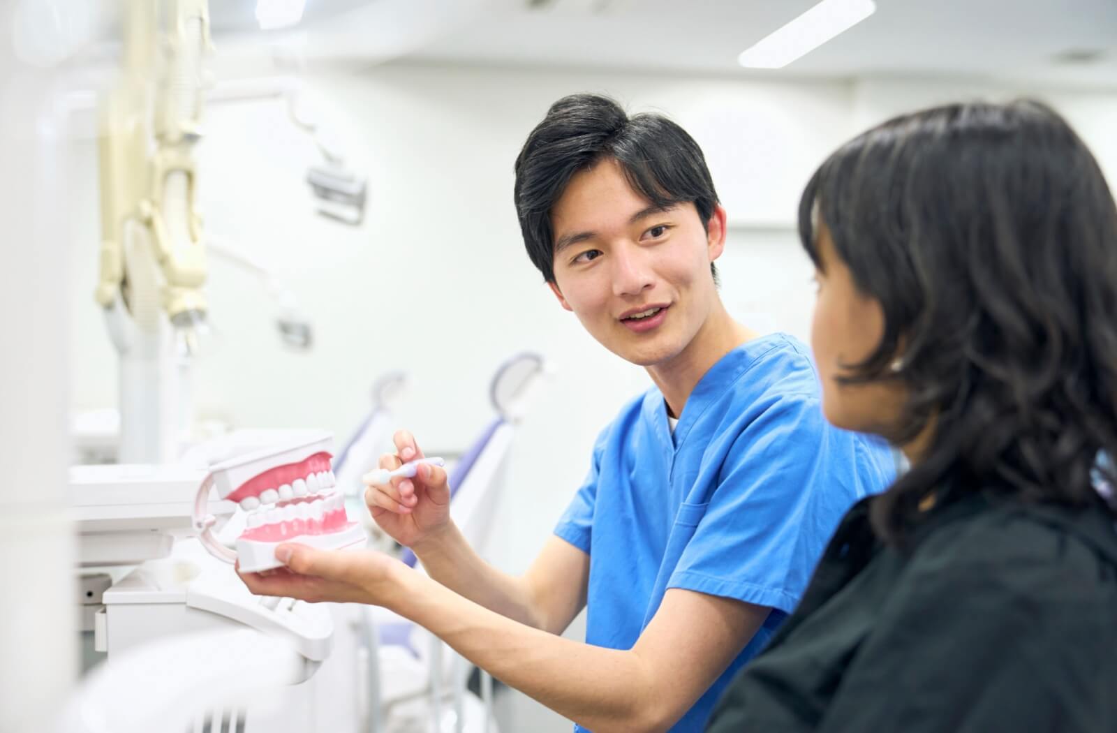 Dentist in blue scrubs explaining proper brushing technique to a patient using a dental model in a modern clinic setting