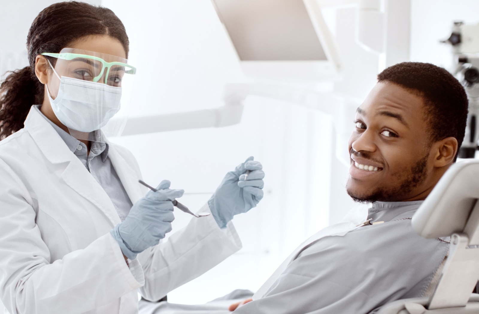 Dental professional wearing a face mask and safety glasses holding dental tools while a patient in a chair smiles confidently