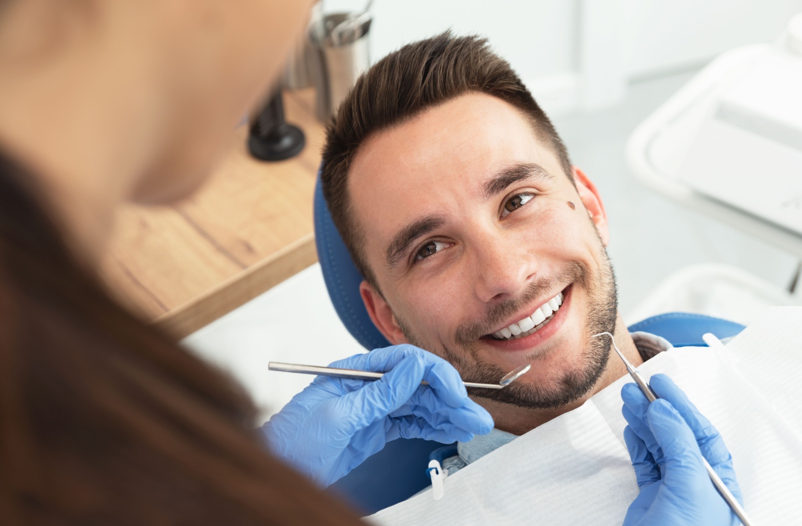 A patient in a dental chair smiling as a dental professional uses tools during an examination